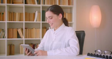 Tableti kullanan genç çekici beyaz iş kadınının dolap portresi ofiste masada otururken mutlu bir şekilde gülüyor