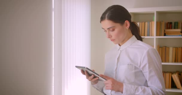 Närbild porträtt av unga attraktiva kaukasiska affärskvinna använder Tablet stående i kontor med bokhyllor på bakgrunden — Stockvideo