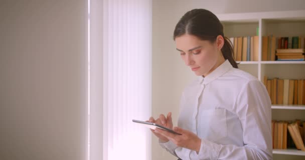Tablet kullanan genç çekici kafkas iş kadınının arka planda kitaplıklarla ofiste duran kameraya bakan yakın çekim portresi — Stok video