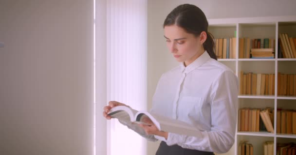 Portrait rapproché de jeune femme d'affaires caucasienne attrayante lisant un livre debout au bureau avec des étagères sur fond — Video