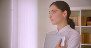 Arka planda ofis raflarında duran kameraya gülümseyen kameraya bakan dizüstü bilgisayar tutan genç kafkas iş kadınının yakın çekim portresi