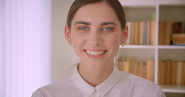 Close-up portret van jonge Kaukasische zakenvrouw kijken naar camera glimlachend gelukkig staande in kantoor met boekenkasten op achtergrond — Stockvideo