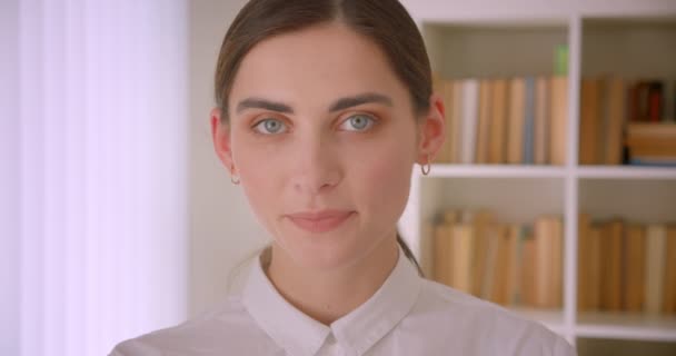Крупный план портрета молодой кавказской предпринимательницы, смотрящей в камеру, радостно улыбающейся стоя в офисе с книжными полками на заднем плане — стоковое видео