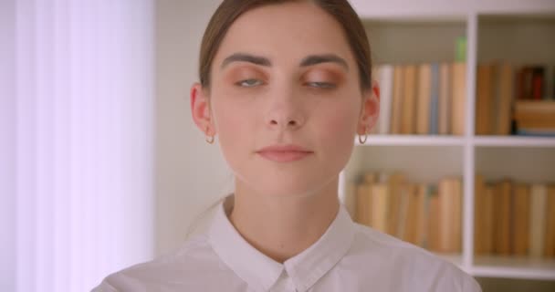 Крупный план портрета молодой кавказской предпринимательницы, смотрящей в камеру, улыбающейся стоя в офисе с книжными полками на заднем плане — стоковое видео
