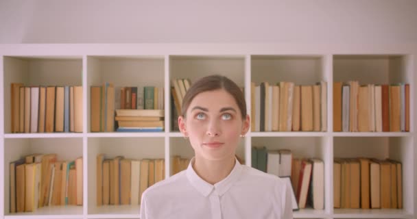 Крупный план портрета молодой веселой кавказской предпринимательницы, смотрящей в камеру, весело улыбающейся стоя на книжных полках офиса на заднем плане — стоковое видео