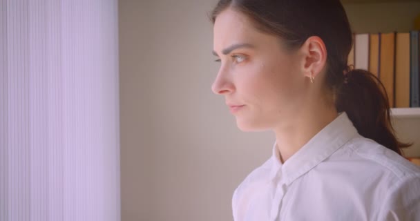 Retrato de cerca de una joven mujer de negocios caucásica mirando por la ventana de pie en la oficina con estanterías en el fondo — Vídeo de stock