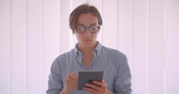 Retrato de primer plano del joven hombre de negocios caucásico guapo usando la tableta de pie en el interior de la oficina — Vídeo de stock