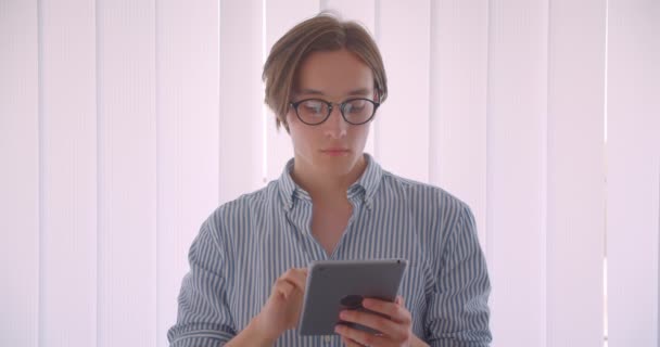 Primer plano retrato de joven guapo hombre de negocios caucásico en gafas usando la tableta de pie en el interior de la oficina — Vídeos de Stock