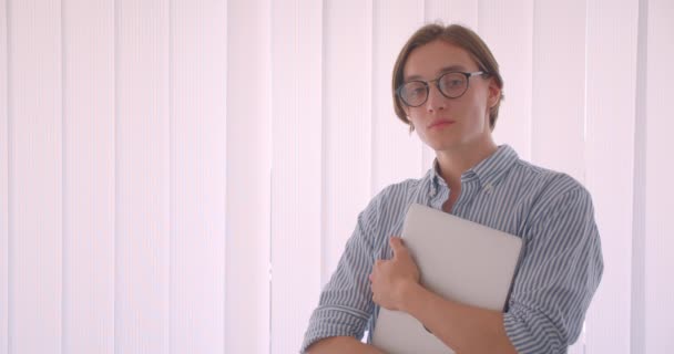 Retrato de close-up do jovem bonito homem de negócios caucasiano em óculos segurando um laptop em pé dentro do escritório — Vídeo de Stock