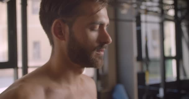 Retrato de close-up de homem caucasiano atraente sem camisa olhando para a frente na frente da câmera e andando no ginásio dentro de casa — Vídeo de Stock