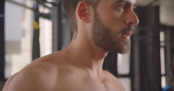 Крупный план портрета мужчины без рубашки и мускулистого кавказца, тренирующегося с гантелями, с усилием стоящего в спортзале в помещении — стоковое видео