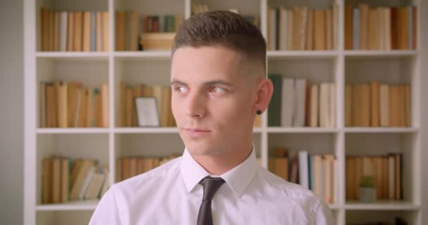 Closeup-portrett av en ung, attraktiv forretningsmann som smiler på biblioteket innendørs med bokhyller på bakgrunn – stockvideo