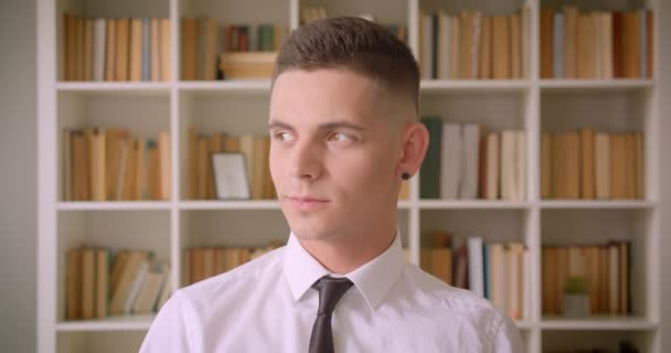 Primo piano ritratto di giovane uomo d'affari attraente guardando la fotocamera sorridente allegramente in biblioteca al chiuso con scaffali su sfondo — Video Stock