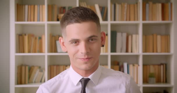 Close-up portret van jonge zelfverzekerde zakenman kijkt naar camera glimlachend gelukkig in bibliotheek binnenshuis met boekenkasten op achtergrond — Stockvideo