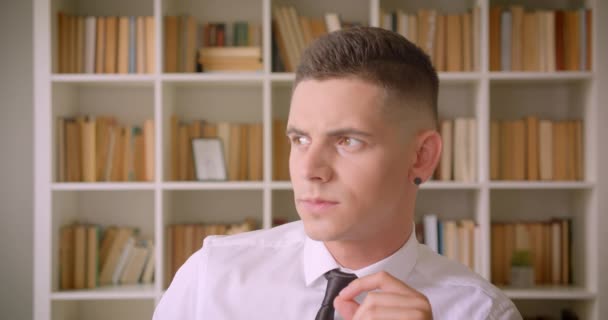 Nahaufnahme Porträt eines jungen erfolgreichen, attraktiven Geschäftsmannes, der in Bibliothek drinnen in die Kamera blickt, mit Bücherregalen im Hintergrund — Stockvideo