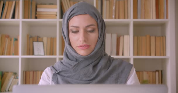 Close-up portret van jonge aantrekkelijke moslim zakenvrouw in hijab met behulp van laptop kijken naar camera glimlachend vrolijk in bibliotheek binnenshuis — Stockvideo