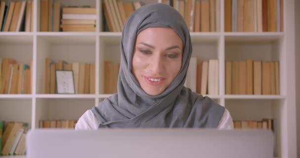 Κοντινό πλάνο του νεαρού ελκυστική μουσουλμανική επιχειρηματίας σε μαντίλα χρησιμοποιώντας φορητό υπολογιστή και να πάρει ενθουσιασμένος στη βιβλιοθήκη σε εσωτερικούς χώρους — Αρχείο Βίντεο