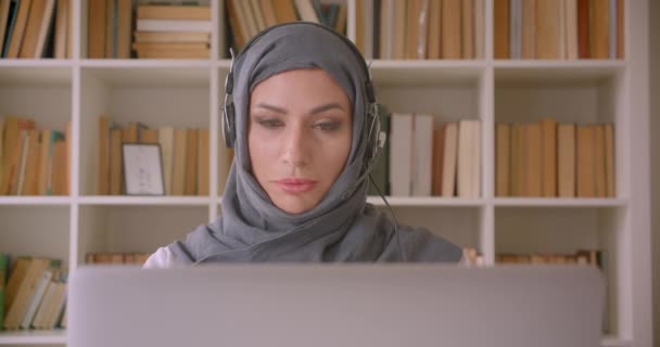 Крупним планом портрет молодої привабливої мусульманської бізнес-леді в хіджабі і навушники з використанням ноутбука і дзвінком в бібліотеці — стокове відео