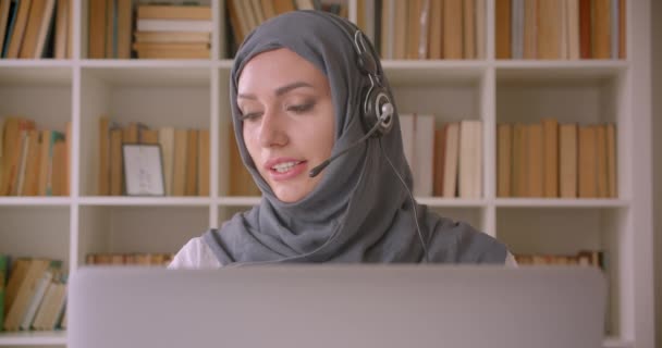 Primo piano ritratto di giovane donna d'affari musulmana attraente in hijab e cuffie utilizzando il computer portatile e avendo una chiamata parlando allegramente in biblioteca — Video Stock