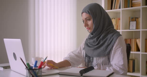 Retrato de cerca de una joven atractiva mujer de negocios musulmana en hijab usando un portátil y tomando notas sentada en el escritorio en la oficina en el interior — Vídeo de stock