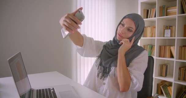 Retrato de cerca de la joven mujer de negocios musulmana en hijab tomando selfies en el teléfono posando sentado frente a la computadora portátil en la oficina en el interior — Vídeo de stock