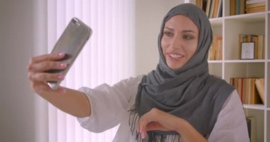 Türbanlı genç neşeli güzel arap iş kadınının, ofiste ki dizüstü bilgisayarın önünde otururken telefonda selfie çektiği closeup portresi