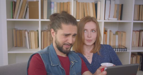 Hipster-Typ spielt Spiel auf Tablet absorbiert und interessiert, während seine Freundin zuschaut und ihn zu Hause unterstützt. — Stockvideo