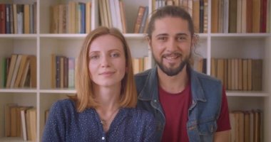 Genç hipster çiftinin kameraya gülümseyip kitaplıklarda birbirlerine sarılarak çekilmiş portresi.