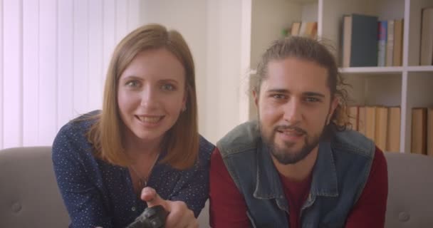 ヒップスター若いカップルは、自宅で1つのチームにあるジョイスティックを使用してビデオゲームをプレイ. — ストック動画