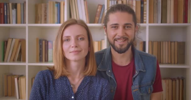 Портрет молодой пары хипстеров, улыбающейся в камеру и обнимающей друг друга на книжных полках . — стоковое видео