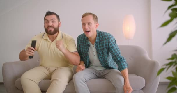 Retrato de close-up de dois homens caucasianos assistindo futebol na TV juntos sentados no sofá em casa dentro de casa — Vídeo de Stock