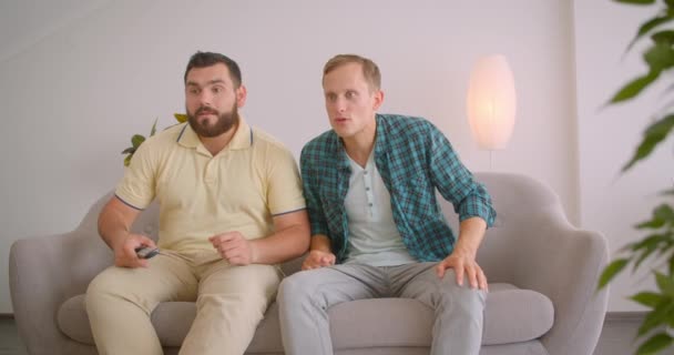 Retrato de close-up de dois homens caucasianos assistindo TV juntos sentados no sofá em casa dentro de casa — Vídeo de Stock