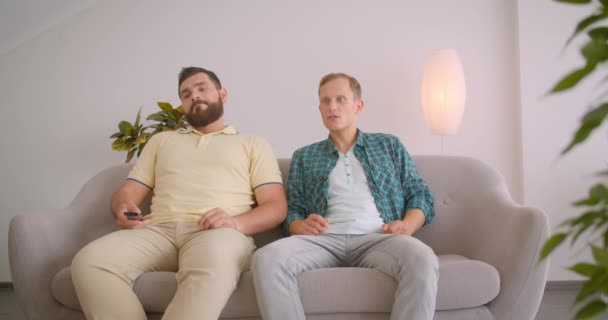 Retrato de cerca de dos hombres caucásicos viendo fútbol en la televisión y celebrando juntos sentados en el sofá en casa dentro — Vídeos de Stock