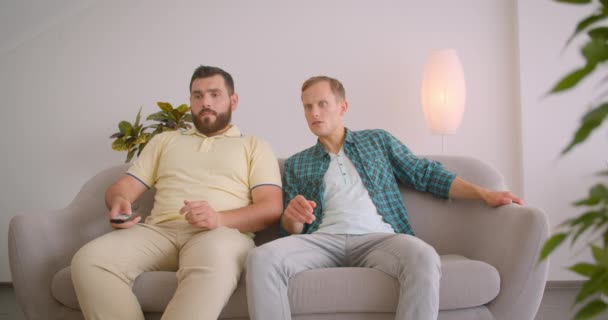 テレビでサッカーを見ている2人の白人男性のクローズアップ肖像画は、自宅の屋内でソファに座って一緒に失われたことに不満を抱いている — ストック動画