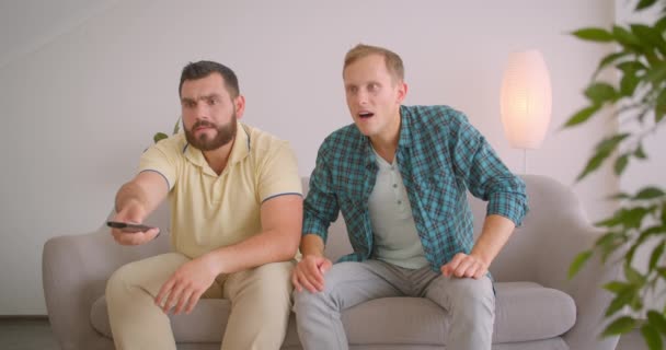 Retrato de close-up de dois adultos caucasianos amigos do sexo masculino assistindo futebol na TV sendo chateado por perdidos juntos sentados no sofá em casa dentro de casa — Vídeo de Stock