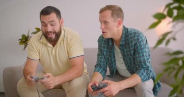 Evde kanepede oturarak birlikte video oyunu oynayan iki yetişkin beyaz adamın yakın çekim portresi. Mutlu adam arkadaşı üzerinde kazanan