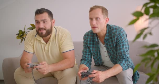 Retrato de cerca de dos hombres caucásicos adultos jugando videojuegos juntos sentados en el sofá en casa dentro. Hombre alegre ganándose a su amigo — Vídeos de Stock
