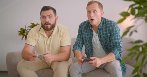 Крупним планом портрет двох кавказьких чоловіків, які грають у відеоігри разом, сидячи на дивані вдома в приміщенні. Веселий чоловік перемагає свого друга — стокове відео