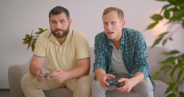 Retrato de cerca de dos hombres adultos jugando videojuegos juntos sentados en el sofá en casa en interiores. Hombre alegre ganándose a su amigo — Vídeo de stock