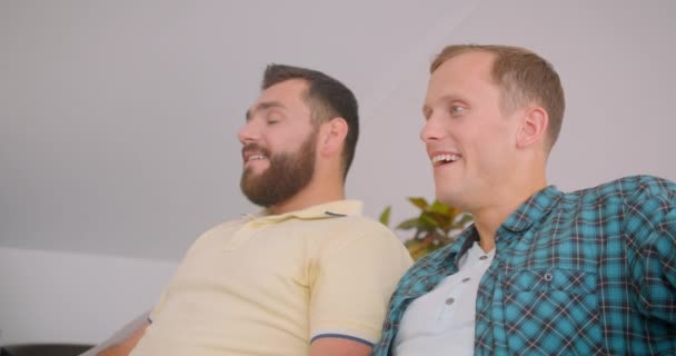 Retrato de close-up de dois homens caucasianos assistindo uma comédia na TV juntos rindo alegremente sentados no sofá em casa dentro de casa — Vídeo de Stock