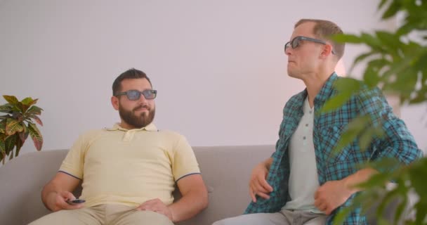 Close-up portret van twee blanke mannen kijken tv samen in 3D bril zittend op de Bank thuis binnenshuis — Stockvideo