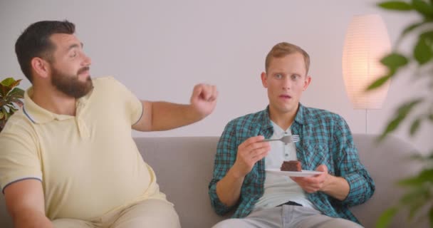 Retrato de cerca de dos hombres caucásicos viendo televisión juntos sentados en el sofá en casa dentro. Hombre triste comiendo pastel de chocolate — Vídeos de Stock