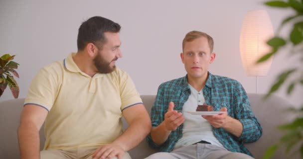 Retrato de cerca de dos hombres caucásicos viendo televisión juntos sentados en el sofá. Hombre deprimido comiendo pastel y siendo apoyado por un amigo — Vídeo de stock