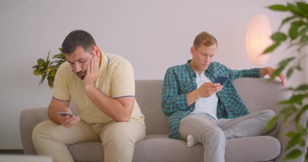 Closeup portrait of two caucasian men using phones sitting on couch together at cozy home indoors — Stock Video