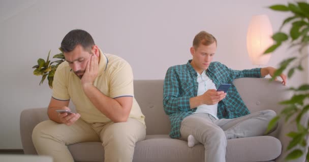 Retrato de perto de dois homens caucasianos usando telefones sentados no sofá juntos dentro de casa — Vídeo de Stock