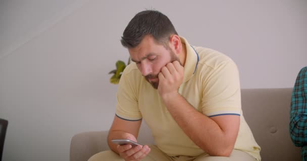 Retrato en movimiento de primer plano de dos hombres caucásicos usando teléfonos sentados en el sofá juntos en el acogedor hogar interior — Vídeo de stock