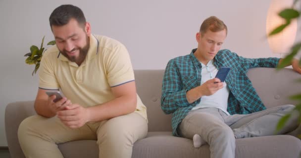 Retrato de cerca de dos hombres caucásicos usando teléfonos sentados en el sofá juntos en el acogedor hogar interior. Amigos compartiendo posts divertidos — Vídeo de stock