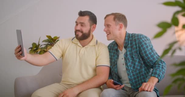 Close-up portret van twee blanke mannen met video-oproep op telefoon zwaaiende Hi zittend op de Bank samen in gezellige huis binnenshuis — Stockvideo