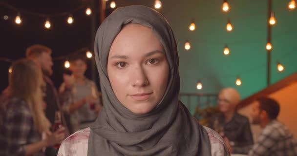Primo piano ritratto di giovane femmina abbastanza musulmana in hijab guardando la fotocamera sorridente allegramente alla festa diversificata in serata accogliente — Video Stock