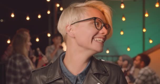 Nahaufnahme Porträt einer jungen hübschen blonden Frau mit Brille, die glücklich in die Kamera lachend auf einer abwechslungsreichen Party am gemütlichen Abend blickt — Stockvideo